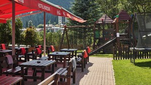 Mobiliario exterior con mesas y bancos, una estructura de juego de madera y un paraguas con la marca Coca-Cola.