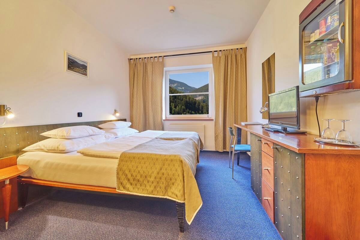 A hotel room with a bed, a desk with a computer and glasses, a window with a view of the outdoors, and a minibar.