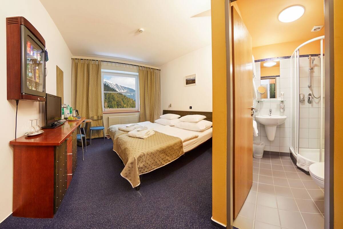 A hotel room with a bed, a desk with a computer, a window with a mountain view, a bathroom with a shower and sink.