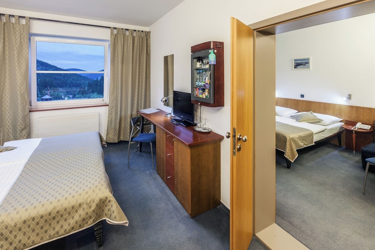 A hotel room with a bed, a desk with a computer, a small fridge, and a window with a view of a forest.