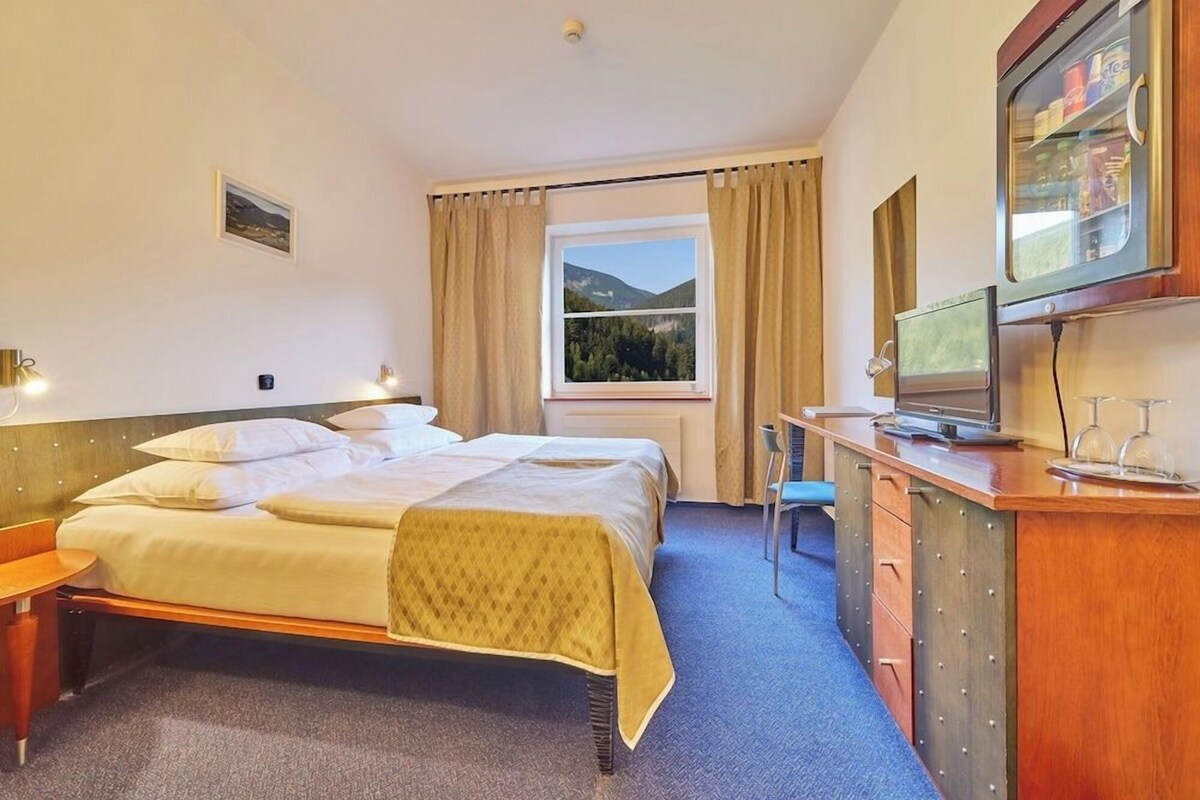 A hotel room with a bed, a desk with a television, a window with a view of mountains, and a small table with a chair.