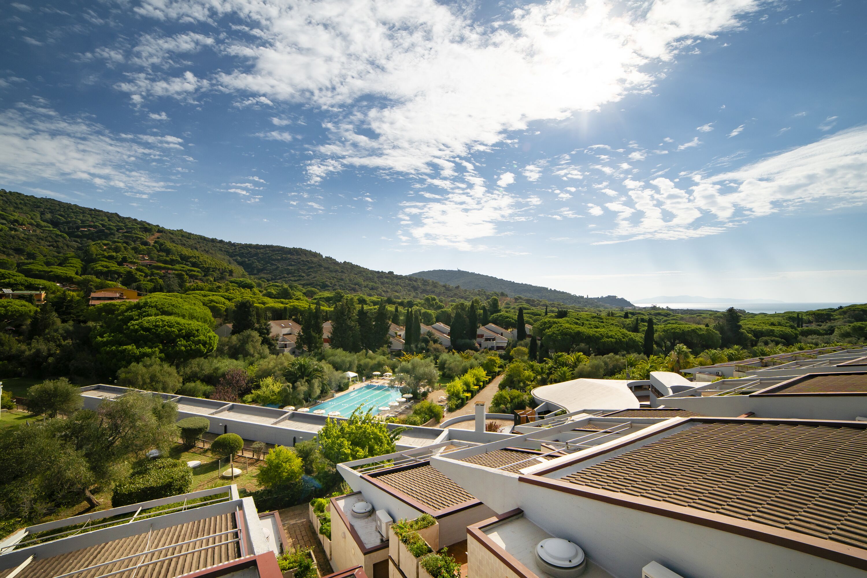 A residential area with a swimming pool, lush greenery, and a hilly landscape in the background.