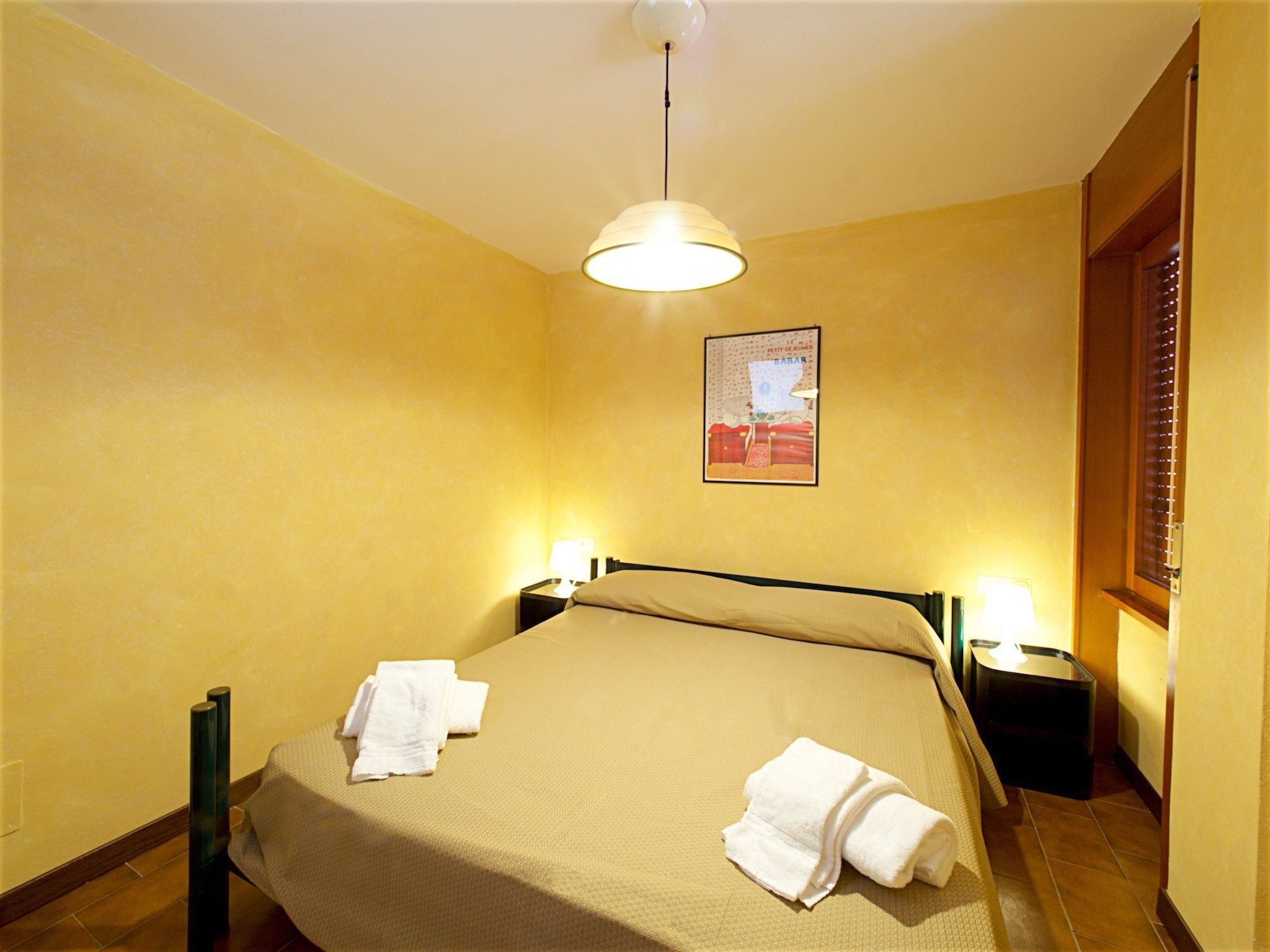 Une chambre d’hôtel avec un lit, deux lampes de chevet, un tableau accroché au mur et une fenêtre avec des stores.