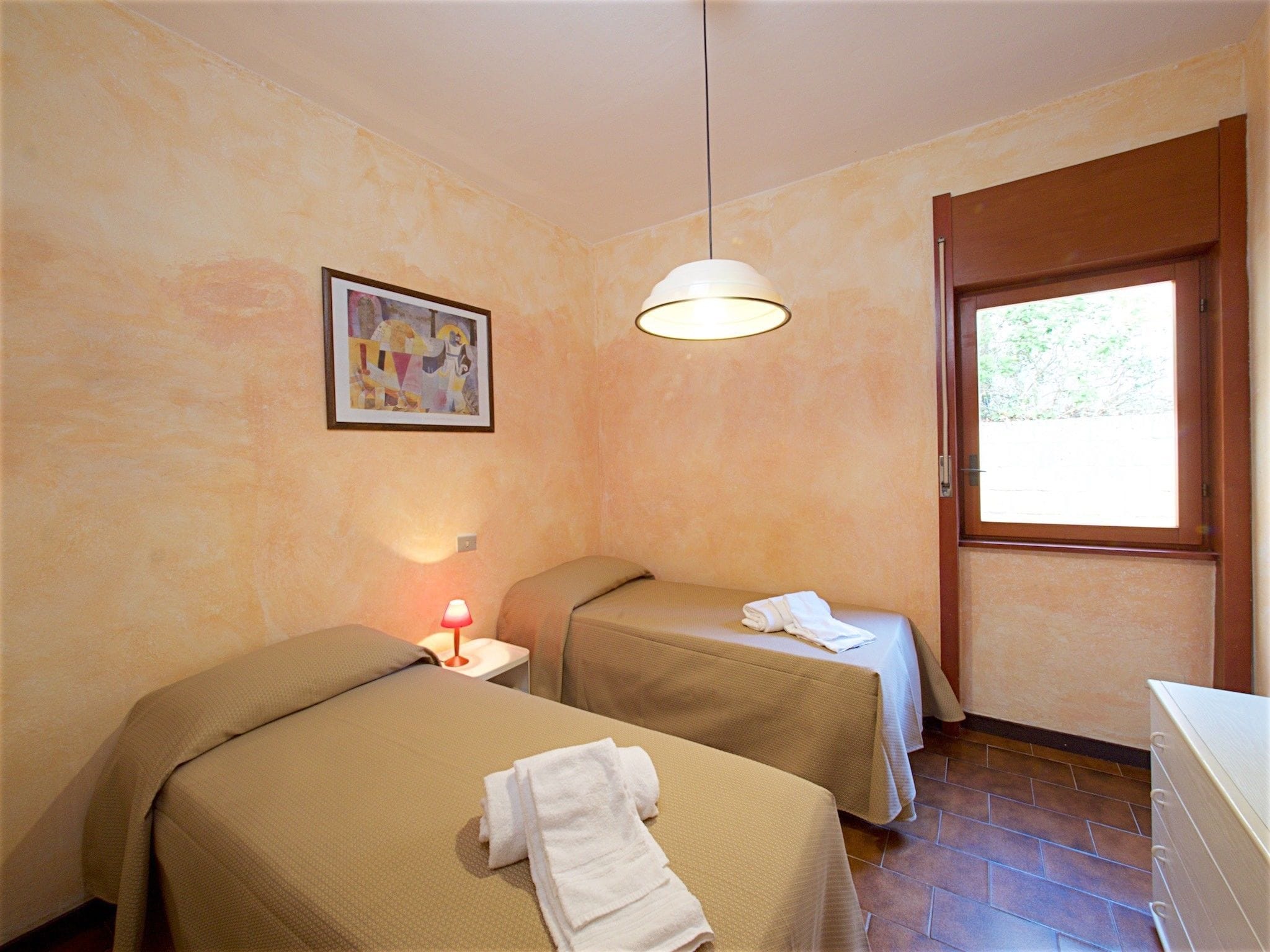 A room with two beds, a painting on the wall, a window, and a lamp.