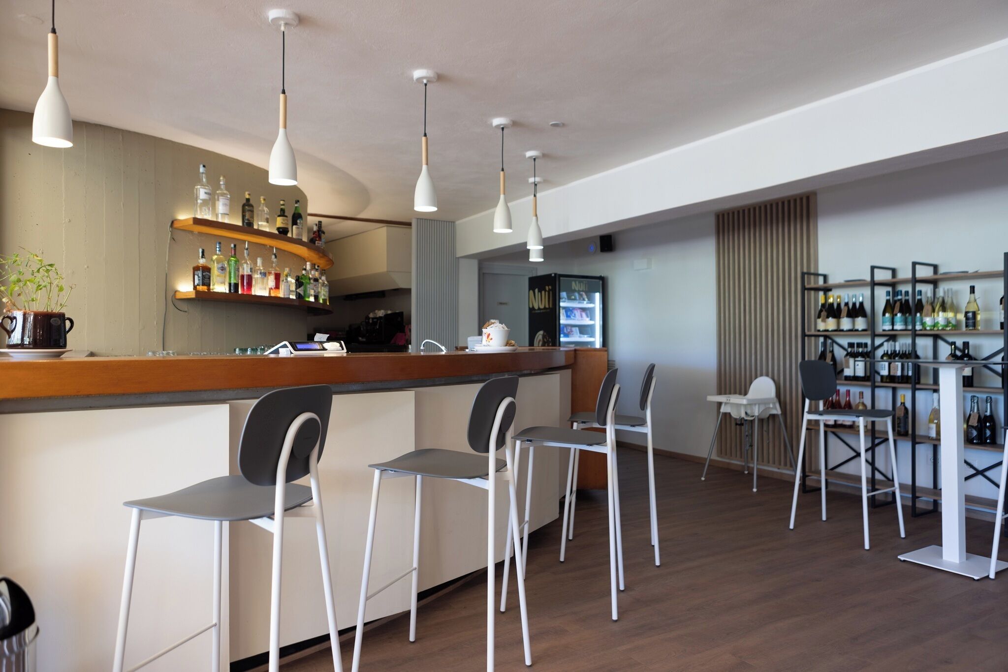 A modern bar area with white bar stools, wooden shelves with bottles, and pendant lighting.