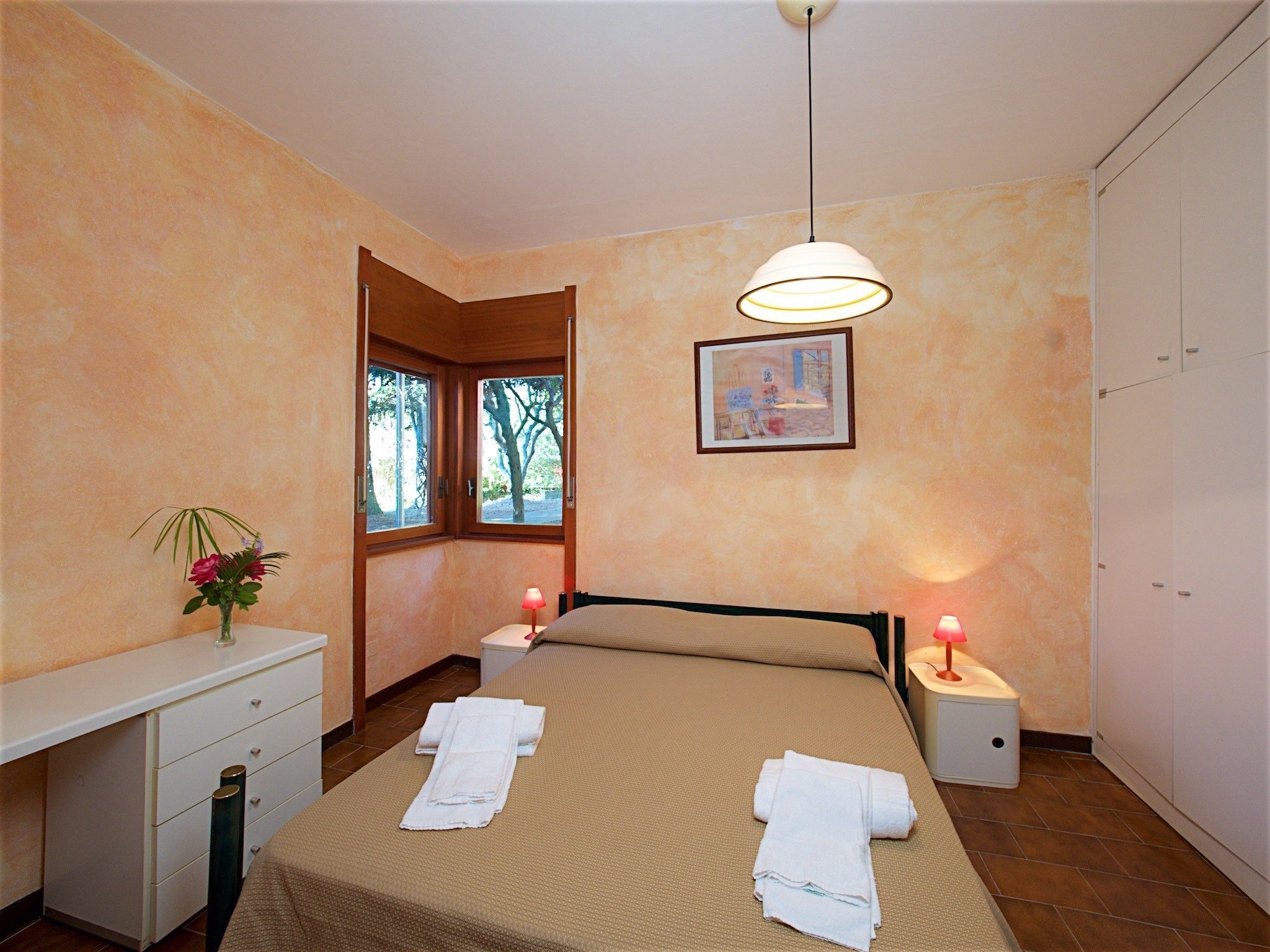 Une chambre à coucher avec un lit, des lampes de chevet, un bureau et une fenêtre donnant sur des arbres.