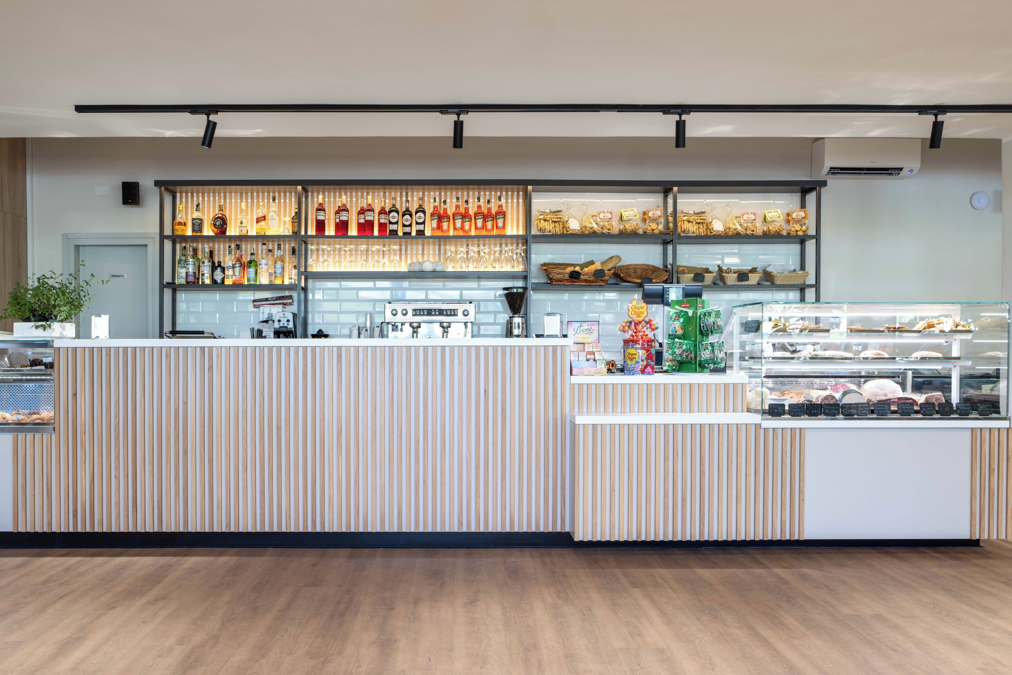 Ett modernt kafé med en bardisk i trä, hyllor fulla av drycker och ett vitrinskåp med bakverk.