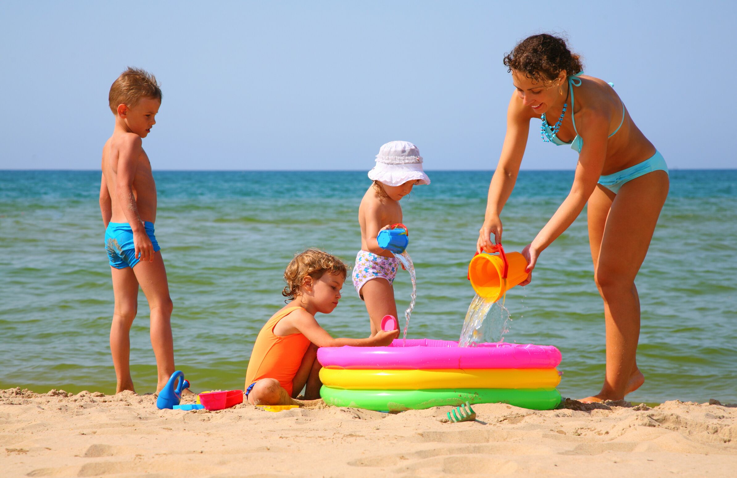 Escena de playa con niños jugando y una mujer llenando un flotador con agua.