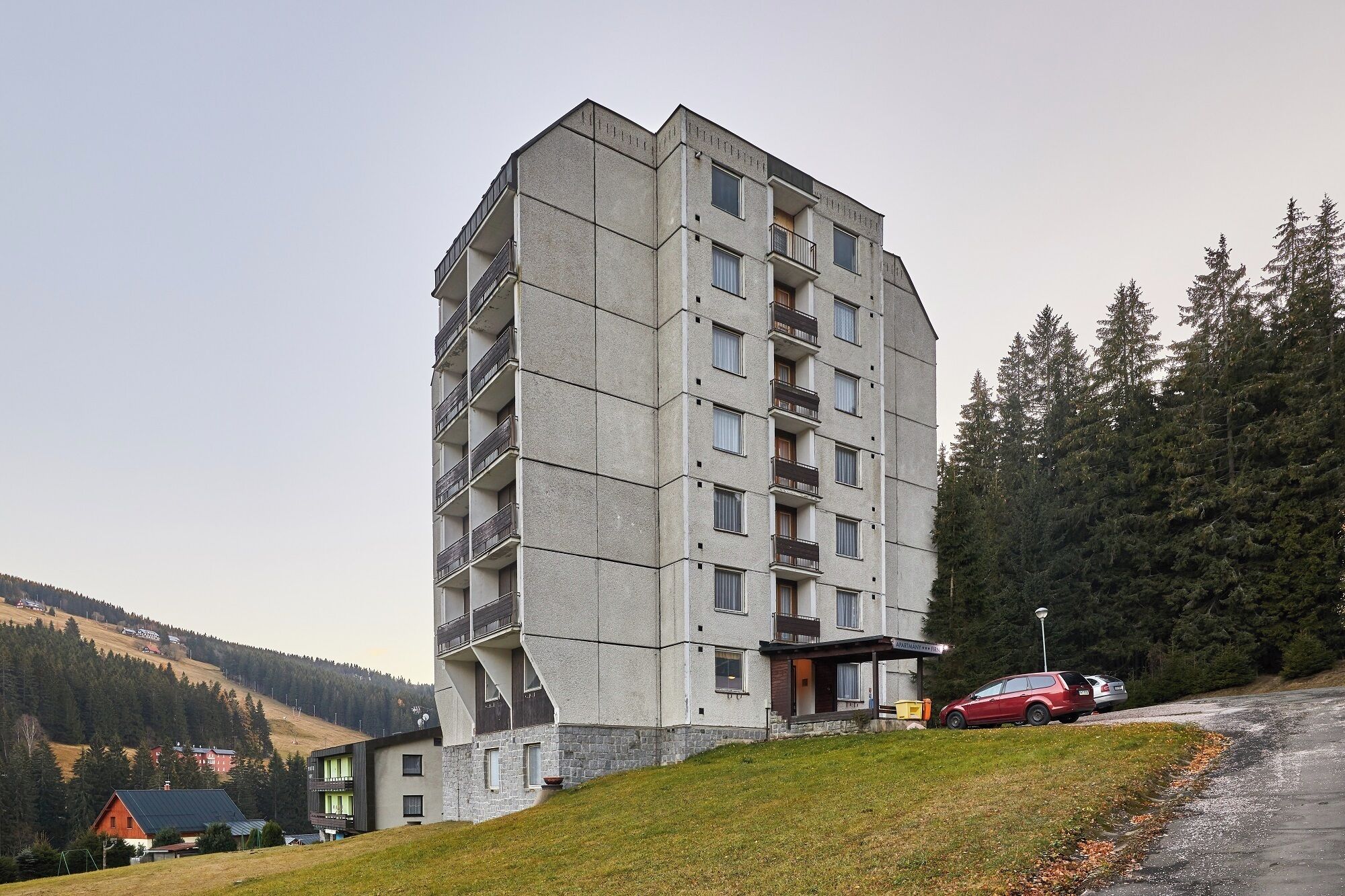 A multi-story building with balconies, a parking area, and a hilly landscape with trees.