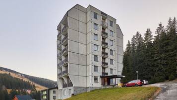 A multi-story building with balconies, a parking area, and a hilly landscape with trees.