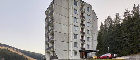 Un edificio de varios pisos con balcones, un estacionamiento y un paisaje montañoso con árboles.
