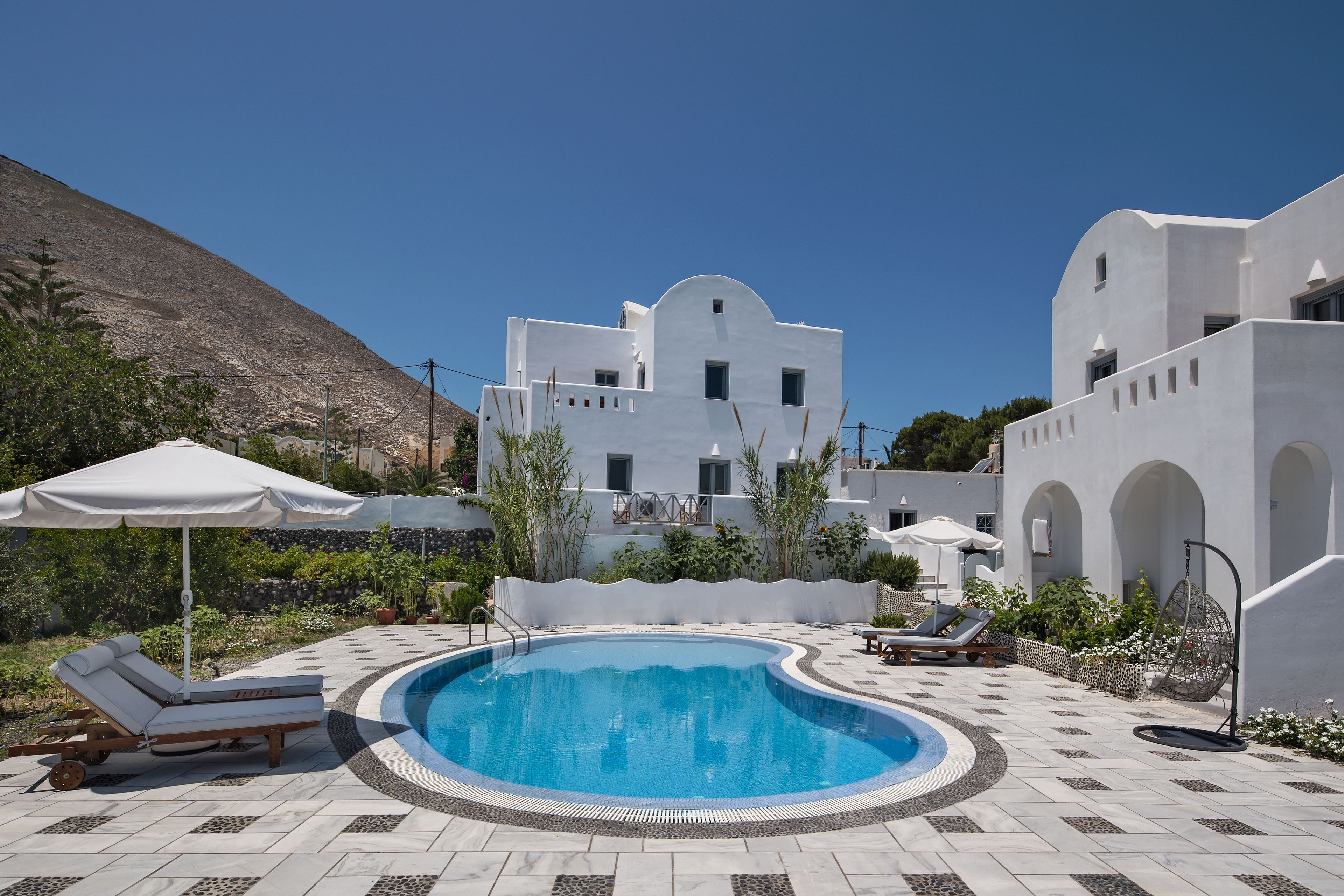 Una piscina con sillones y sombrillas, rodeada de edificios blancos con arcos y montañas al fondo.