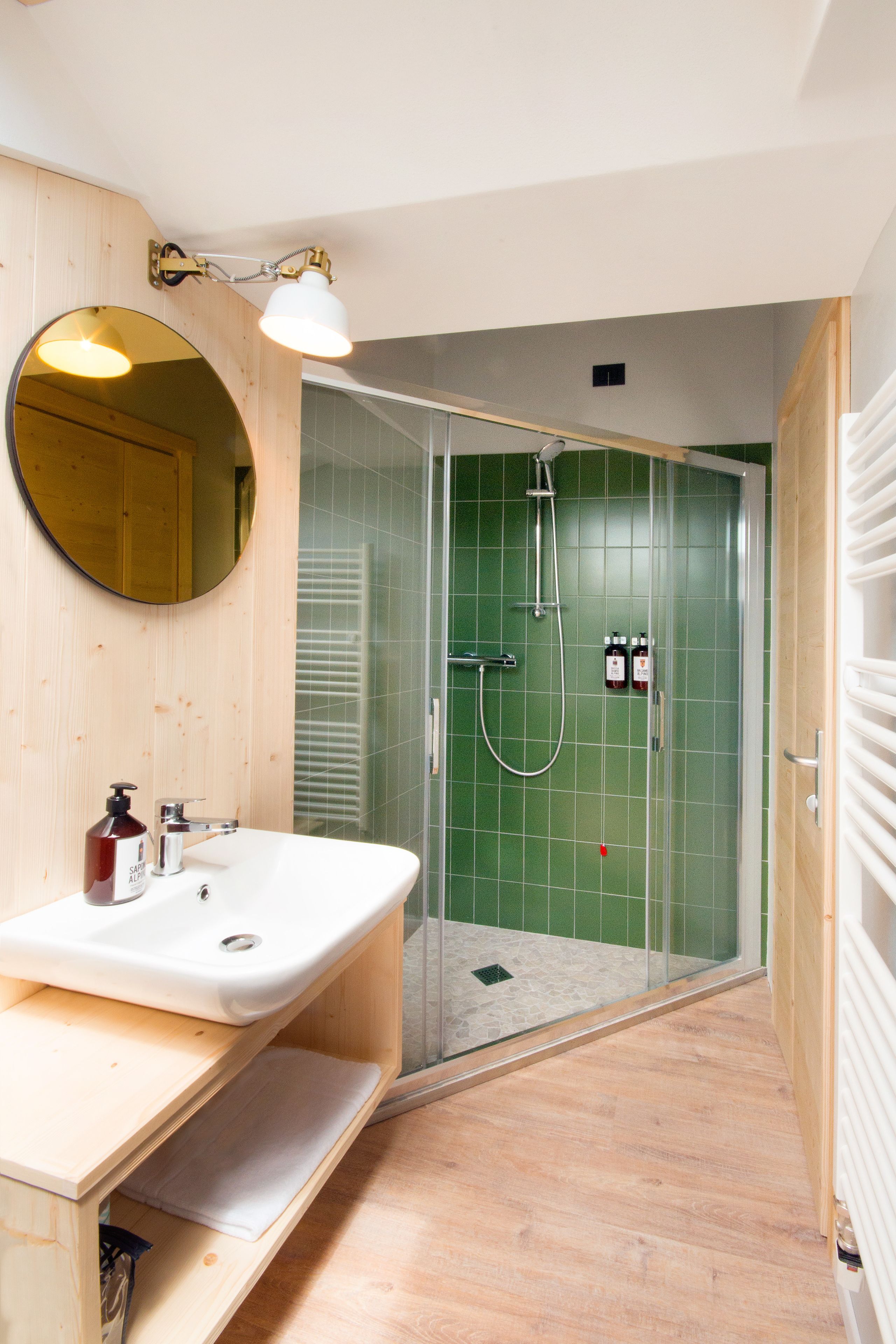 A bathroom with a wooden vanity, a round mirror, a shower with green tiles, and a white radiator.