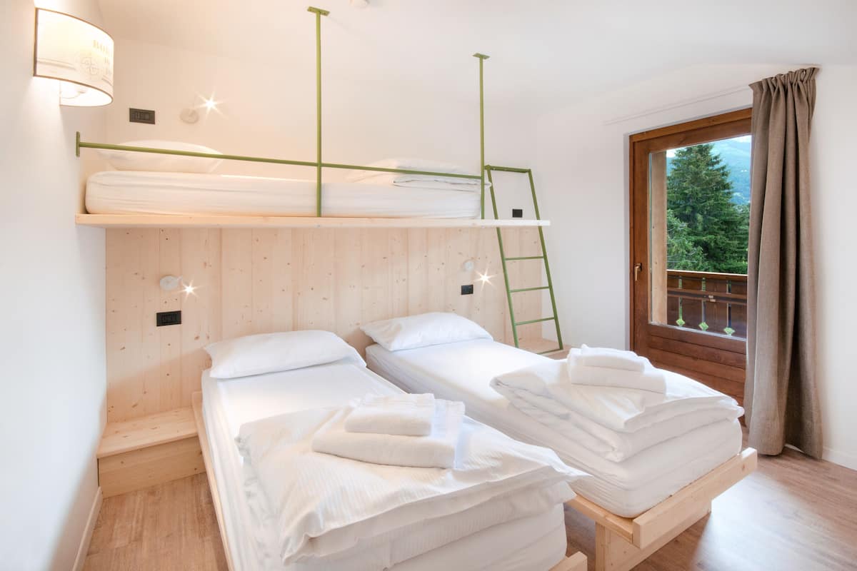 A room with two single beds, a bunk bed, wooden walls, a window with a view of trees, and a ladder.