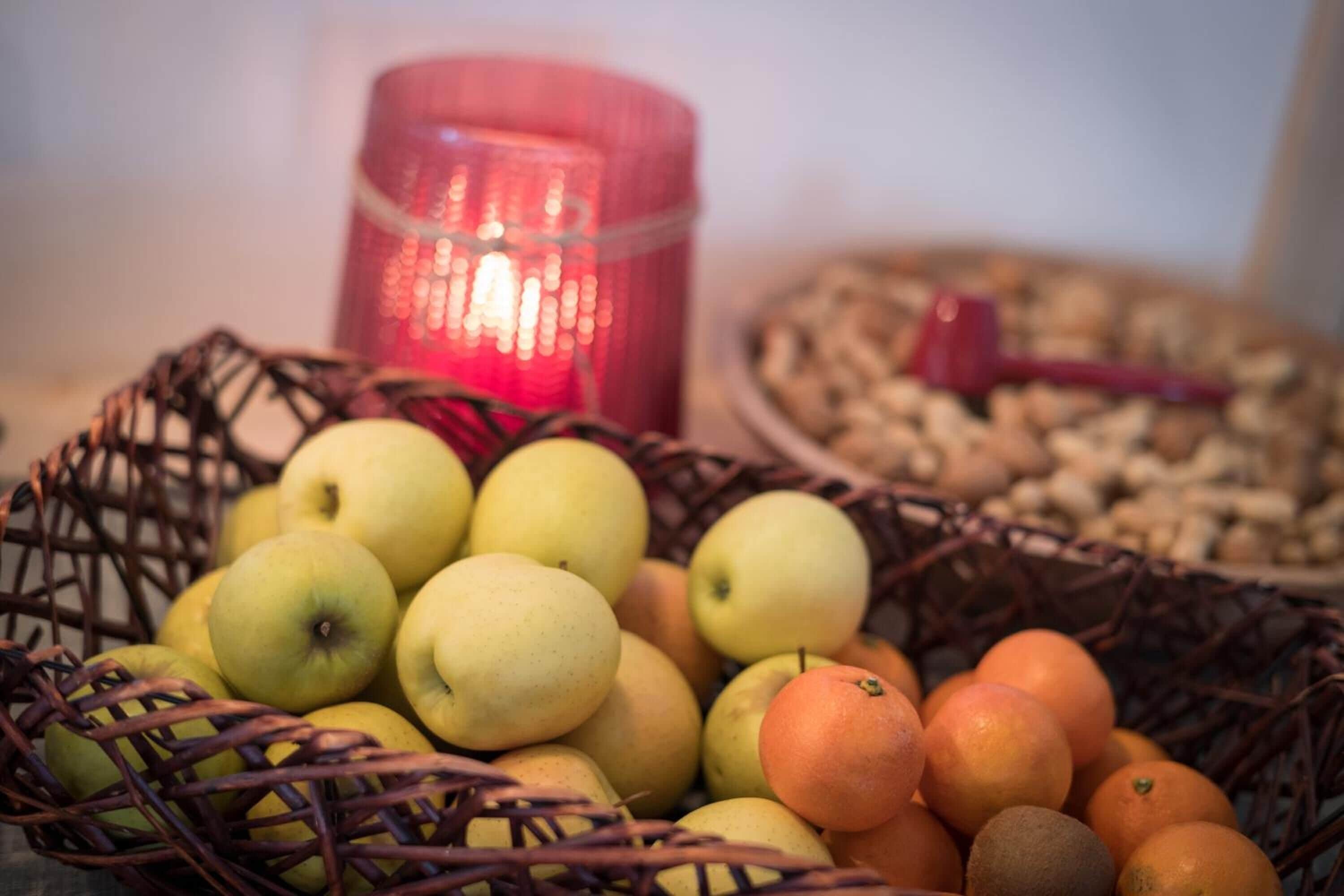 Una cesta de mimbre con manzanas y naranjas, una vela roja y un cuenco de nueces al fondo.