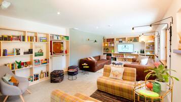 Una sala de estar acogedora con un sofá, sillones y una mesa de centro. Hay estanterías llenas de libros y objetos decorativos. Un televisor está montado en la pared, y se ve un radiador.