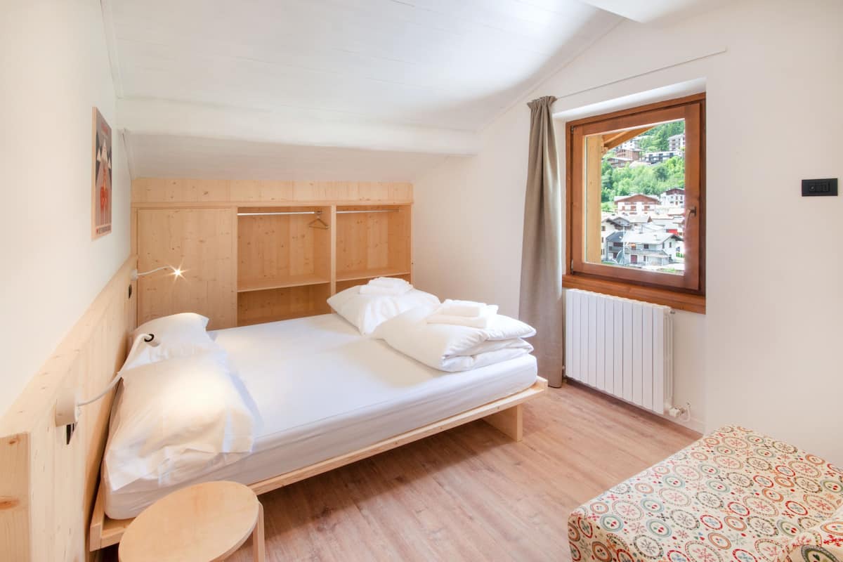 A single bed with a wooden headboard, a small wooden stool, a window with a view of a town, and a radiator.