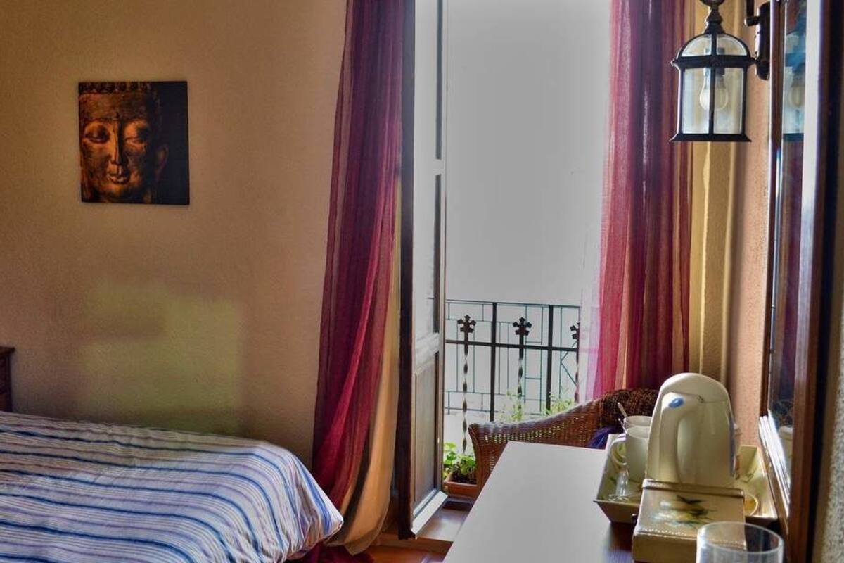 Un dormitorio con cama, mesita de noche, lámpara, ventana con cortinas y vista a un balcón.