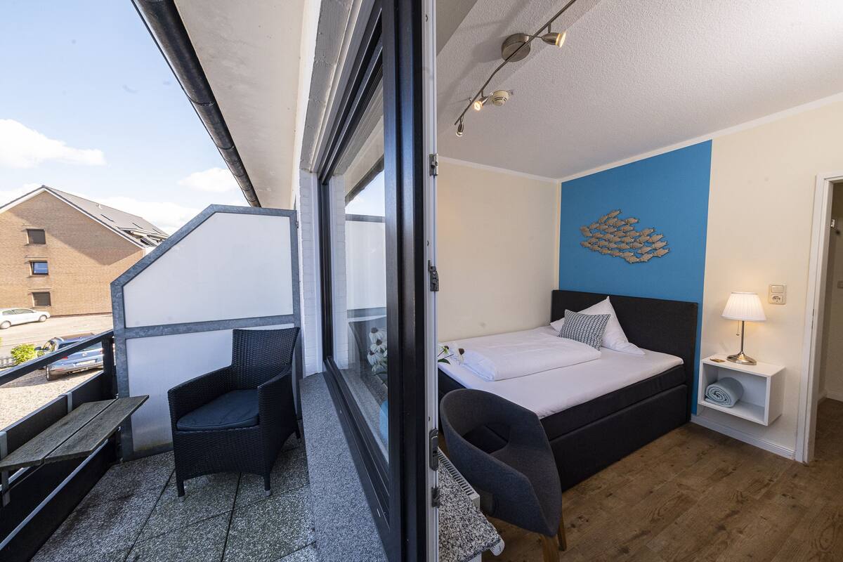 A hotel room with a bed, a chair, a small table, a nightstand with a lamp, and a view of the outside.