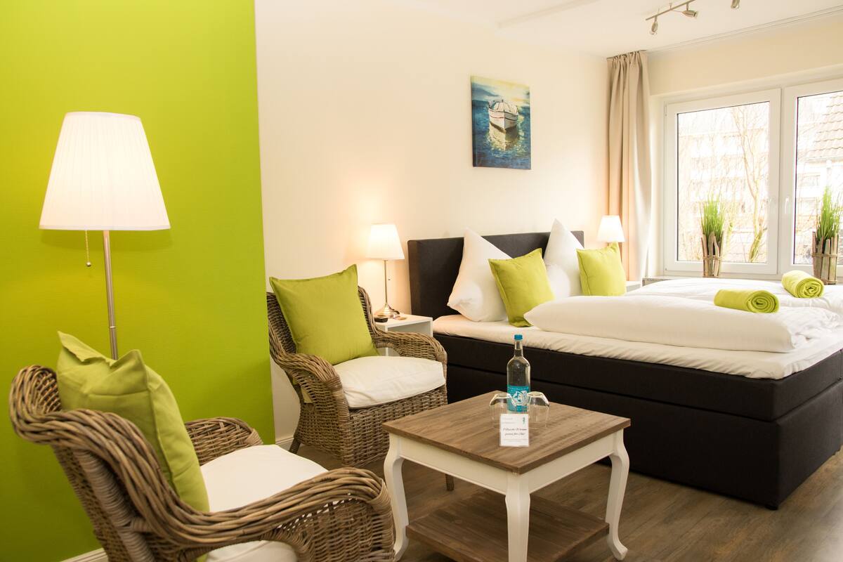 A hotel room with a bed, two wicker chairs with cushions, a small table with a bottle and a sign, and a window with plants.