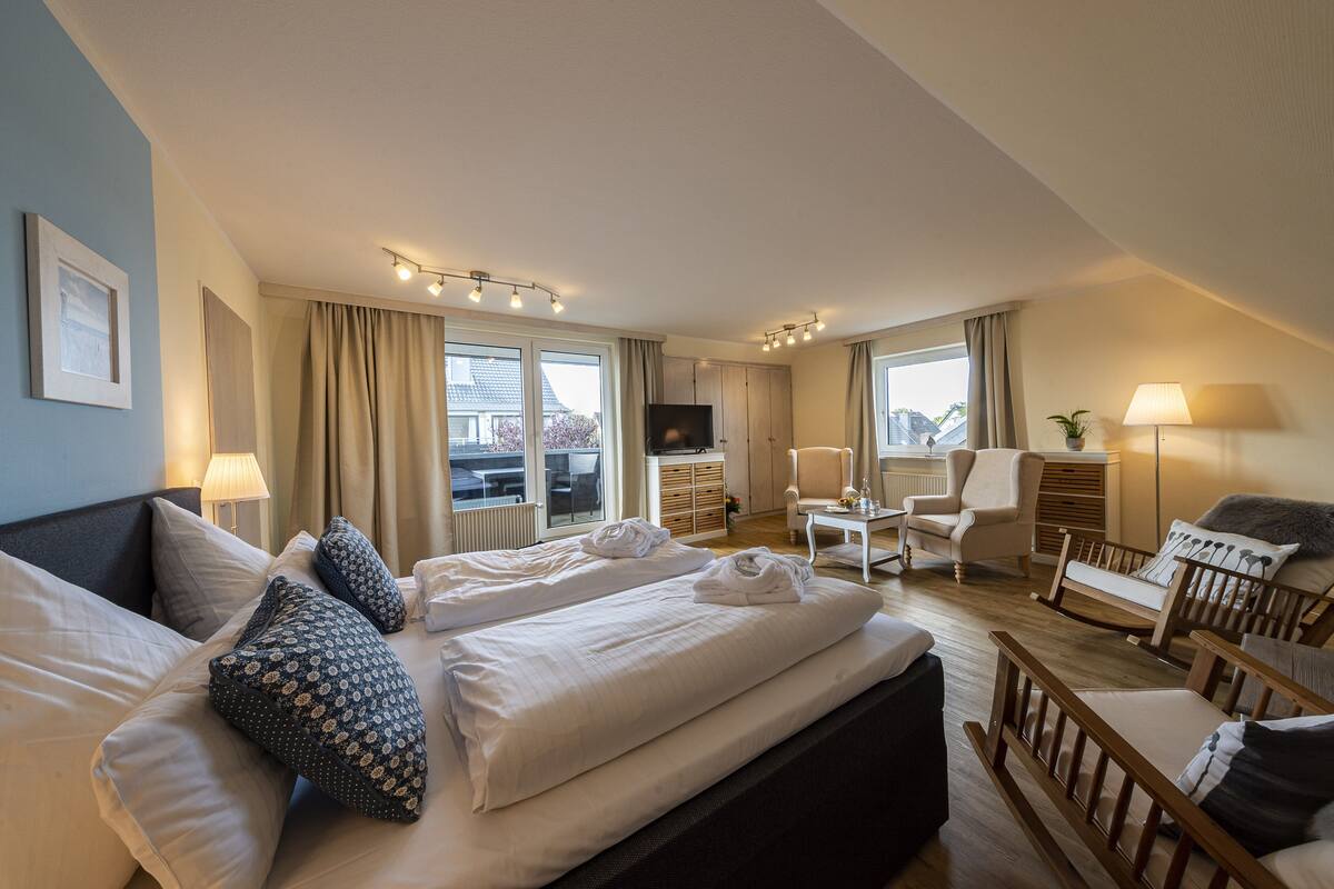 A hotel room with a large bed, two armchairs, a small table, a TV, and a window with curtains.