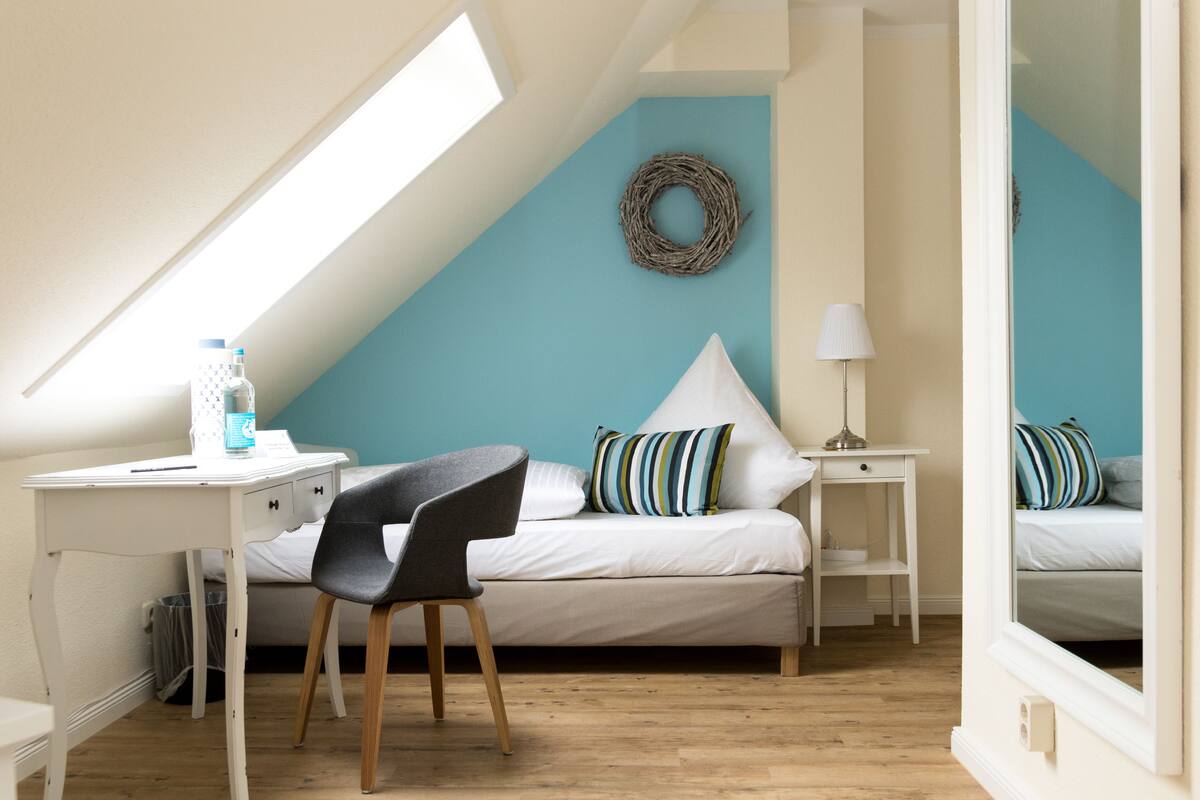 A bedroom with a sloped ceiling, a bed with striped pillows, a desk with a chair, and a small table with a lamp.