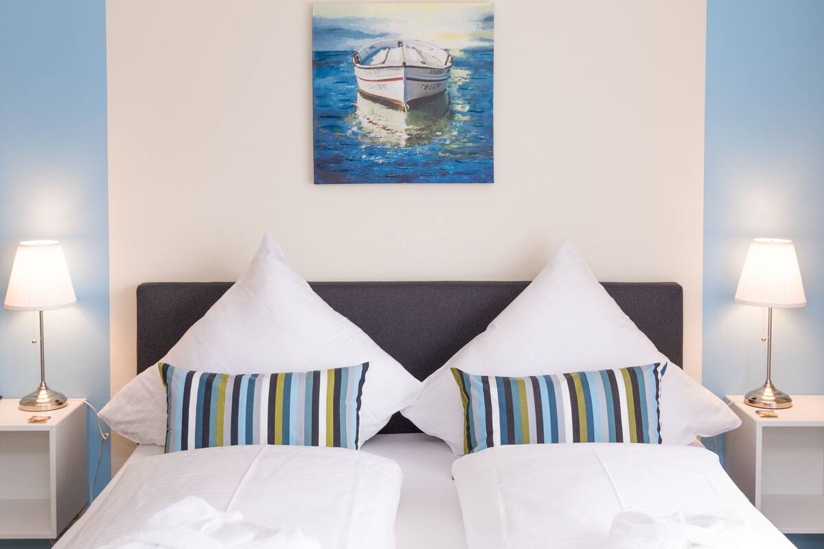 A hotel room with a bed, two pillows, a painting of a boat, and two bedside lamps.