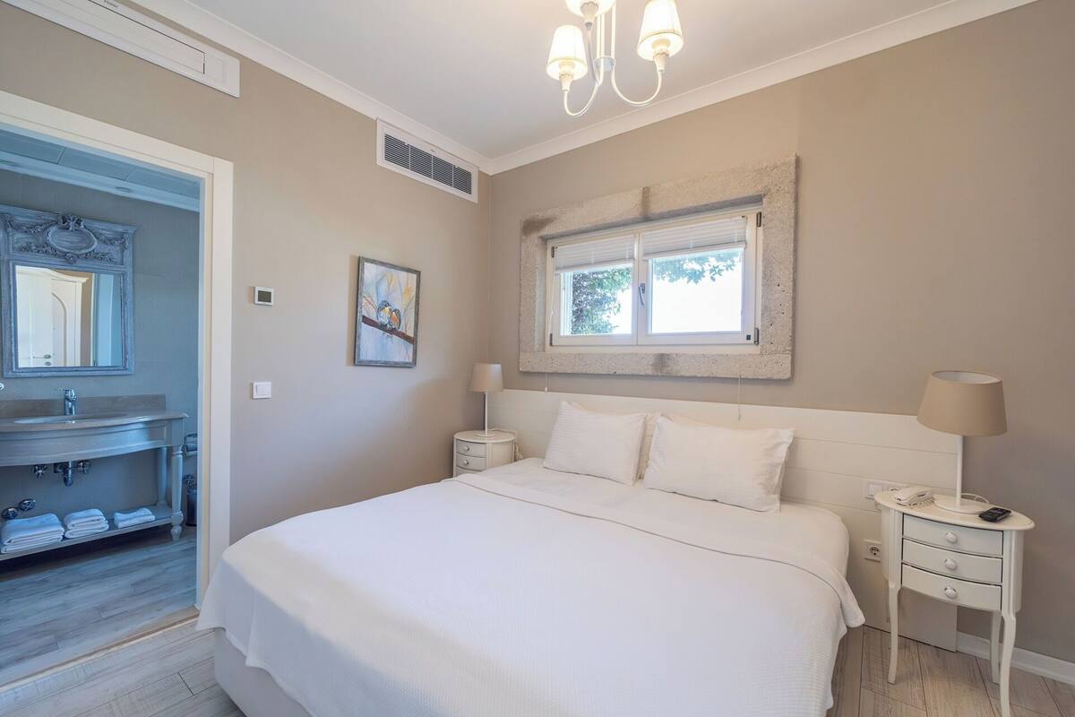 A bedroom with a bed, bedside tables, a nightstand, a window, a mirror, a bathroom with a sink, and a towel rack.