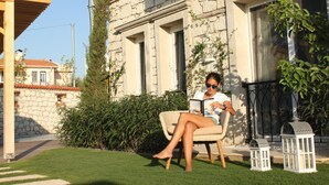 A person sitting on a wicker chair in a garden area with a stone building in the background.