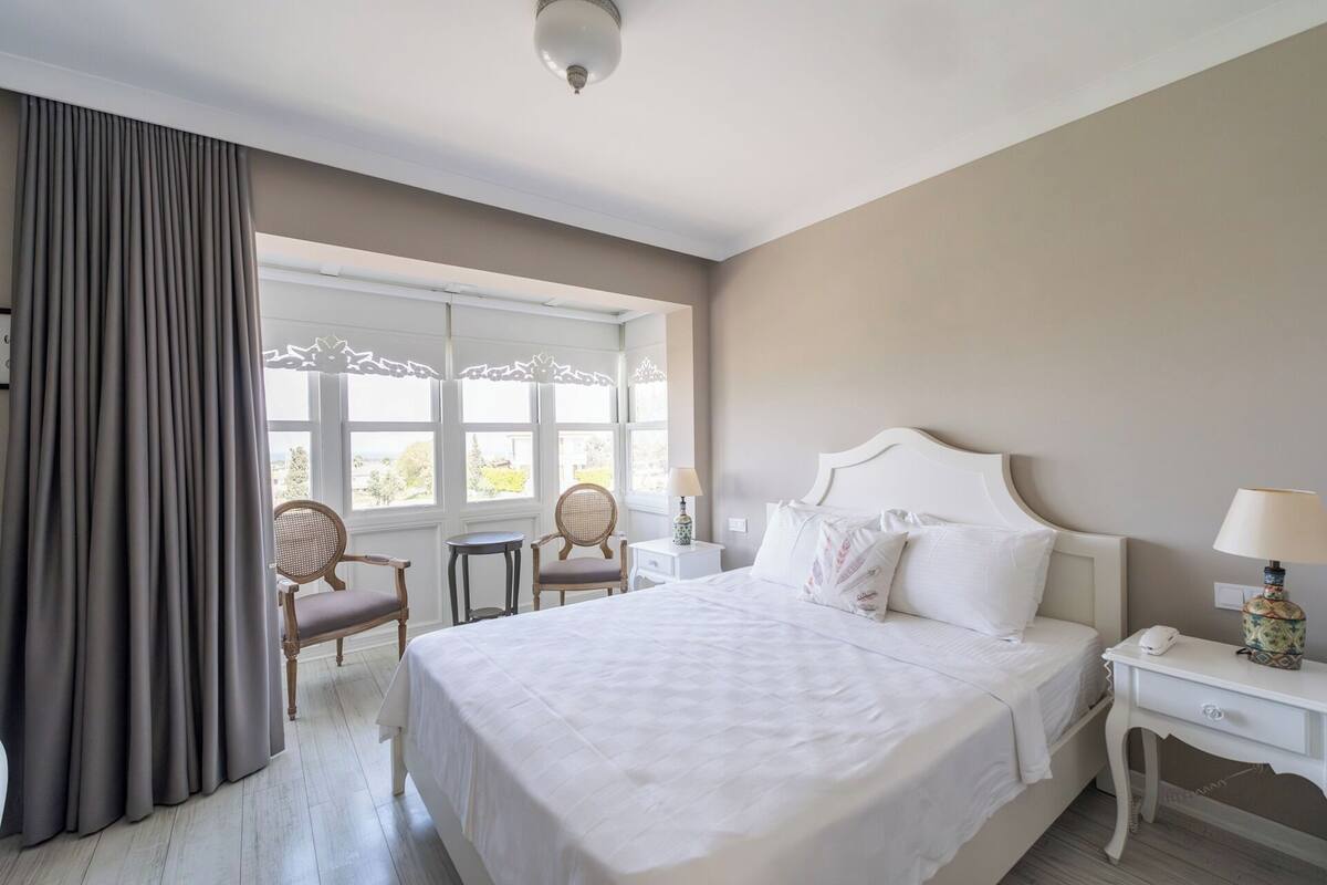 A hotel room with a large bed, a bedside table with a lamp, a chair, and a window with curtains.