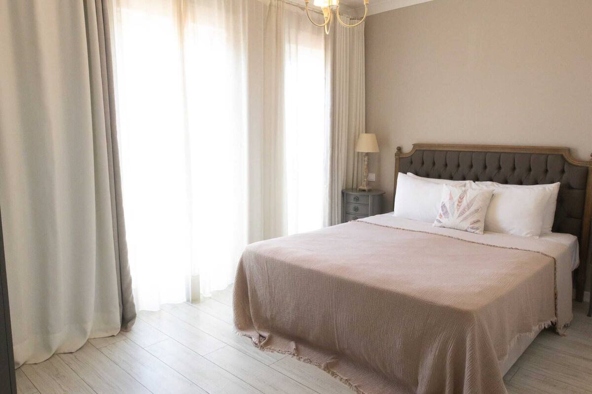 A neatly made bed with a headboard, a bedside table with a lamp, and sheer curtains on a window.