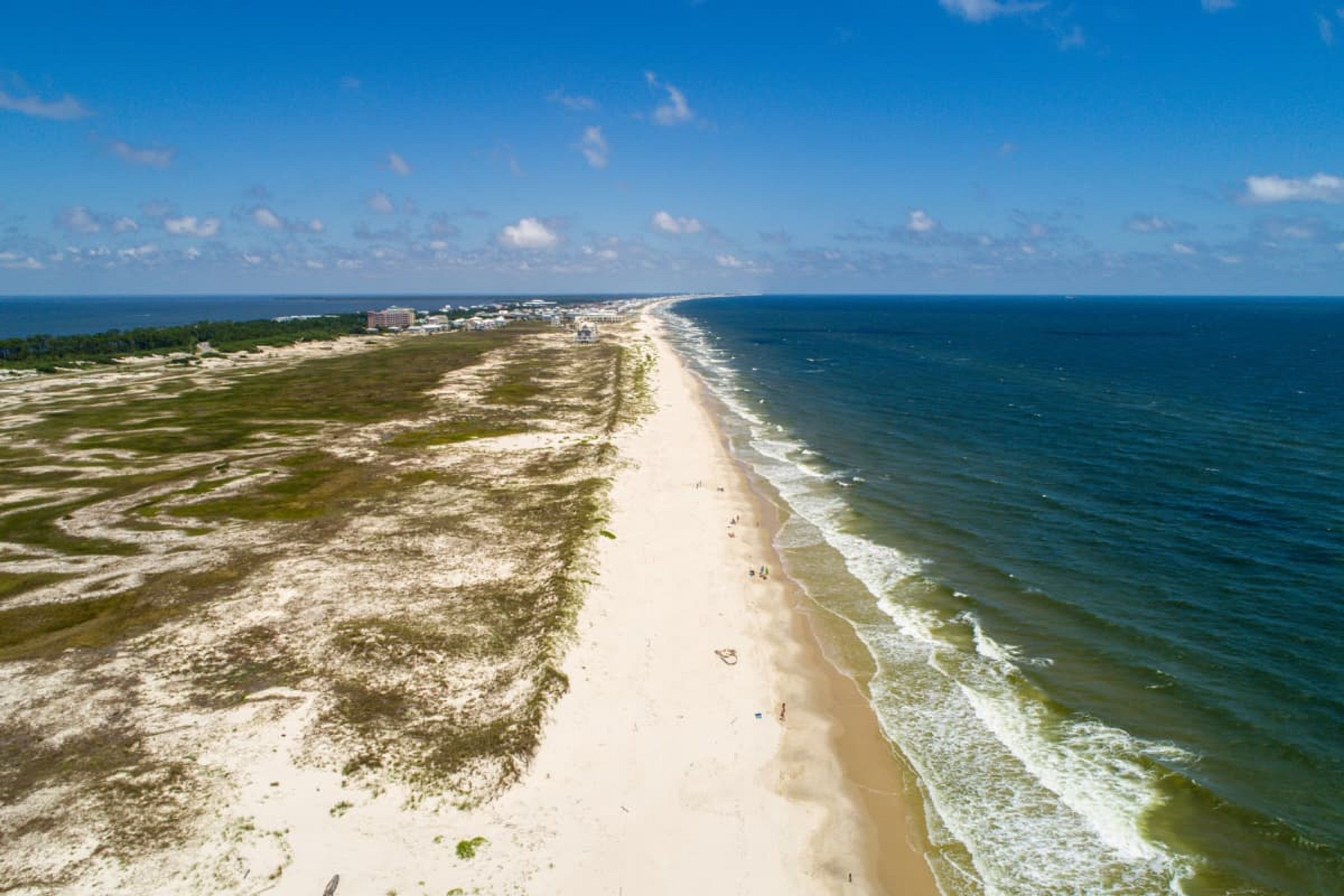 An aerial view of a beach with a long stretch of sand, a few buildings in the distance, and the ocean on one side.