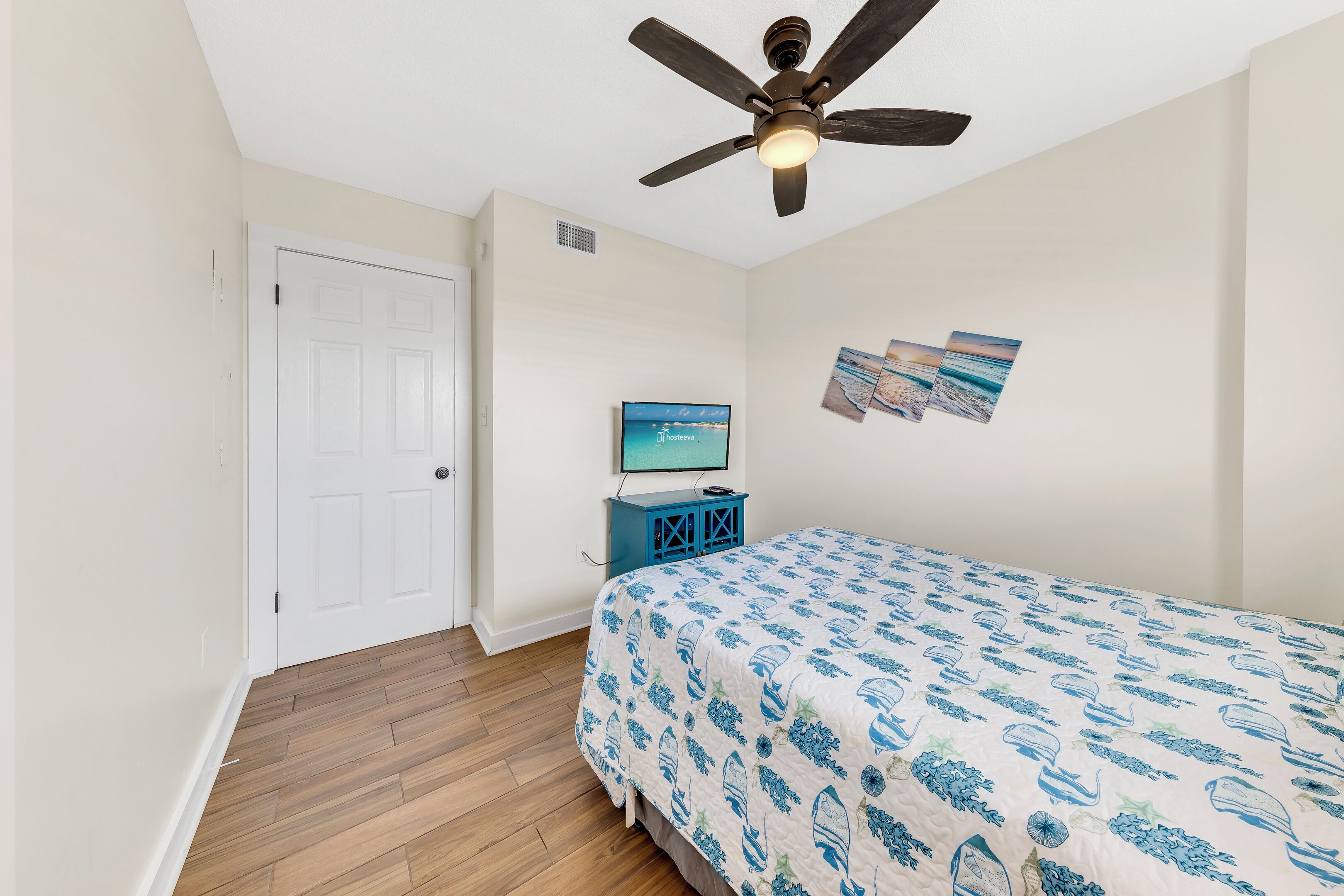 Une chambre à coucher avec un lit, un ventilateur de plafond, un téléviseur posé sur un meuble et une porte.