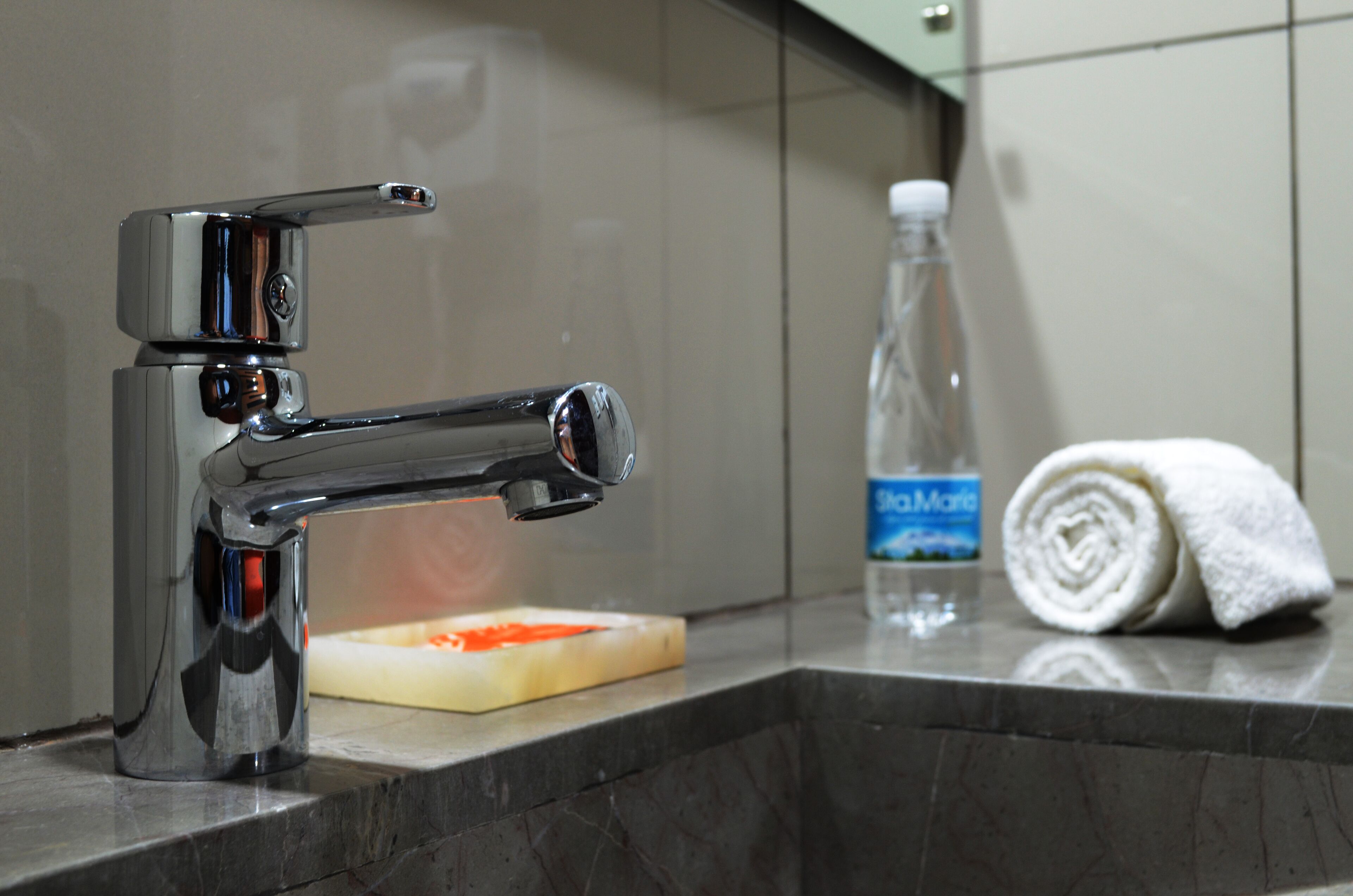 A modern bathroom with a chrome faucet, a soap dish, a bottle of water, and a rolled towel on the counter.