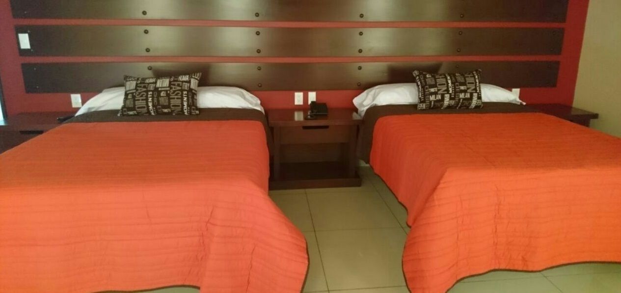 Habitación doble con dos camas, cada una con colcha roja y almohadas a juego. Un mesita de noche entre las camas presenta una lámpara y una pequeña mesa.