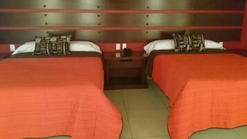 Habitación doble con dos camas, cada una con colcha roja y almohadas a juego. Un mesita de noche entre las camas presenta una lámpara y una pequeña mesa.