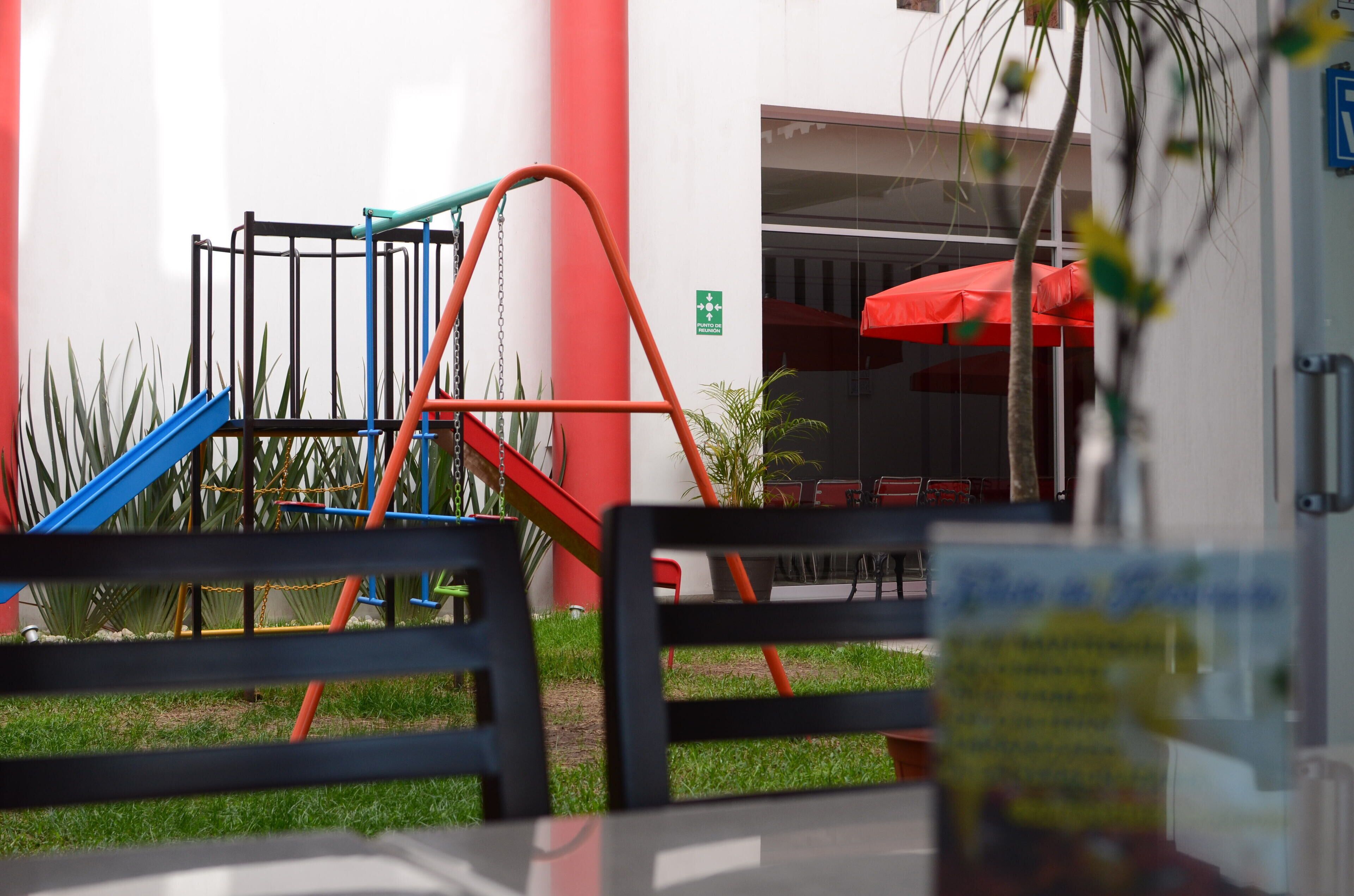 Un área de juegos al aire libre con tobogán y estructura para escalar, un paraguas rojo y una zona de estar con mesas y sillas.