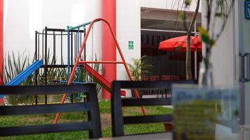 Un área de juegos al aire libre con tobogán y estructura para escalar, un paraguas rojo y una zona de estar con mesas y sillas.
