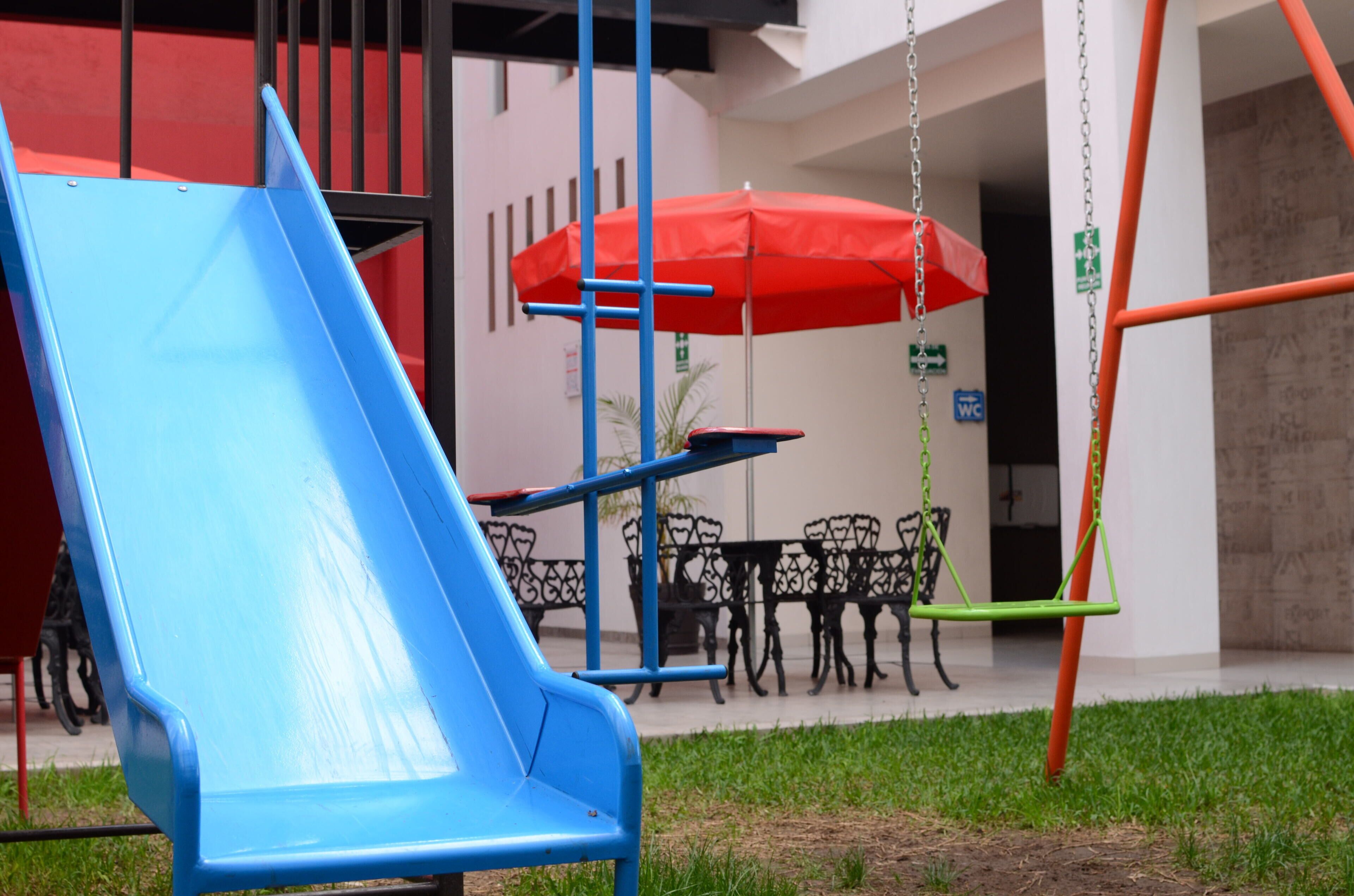 Un parque infantil con un tobogán azul, una sombrilla roja y columpios.