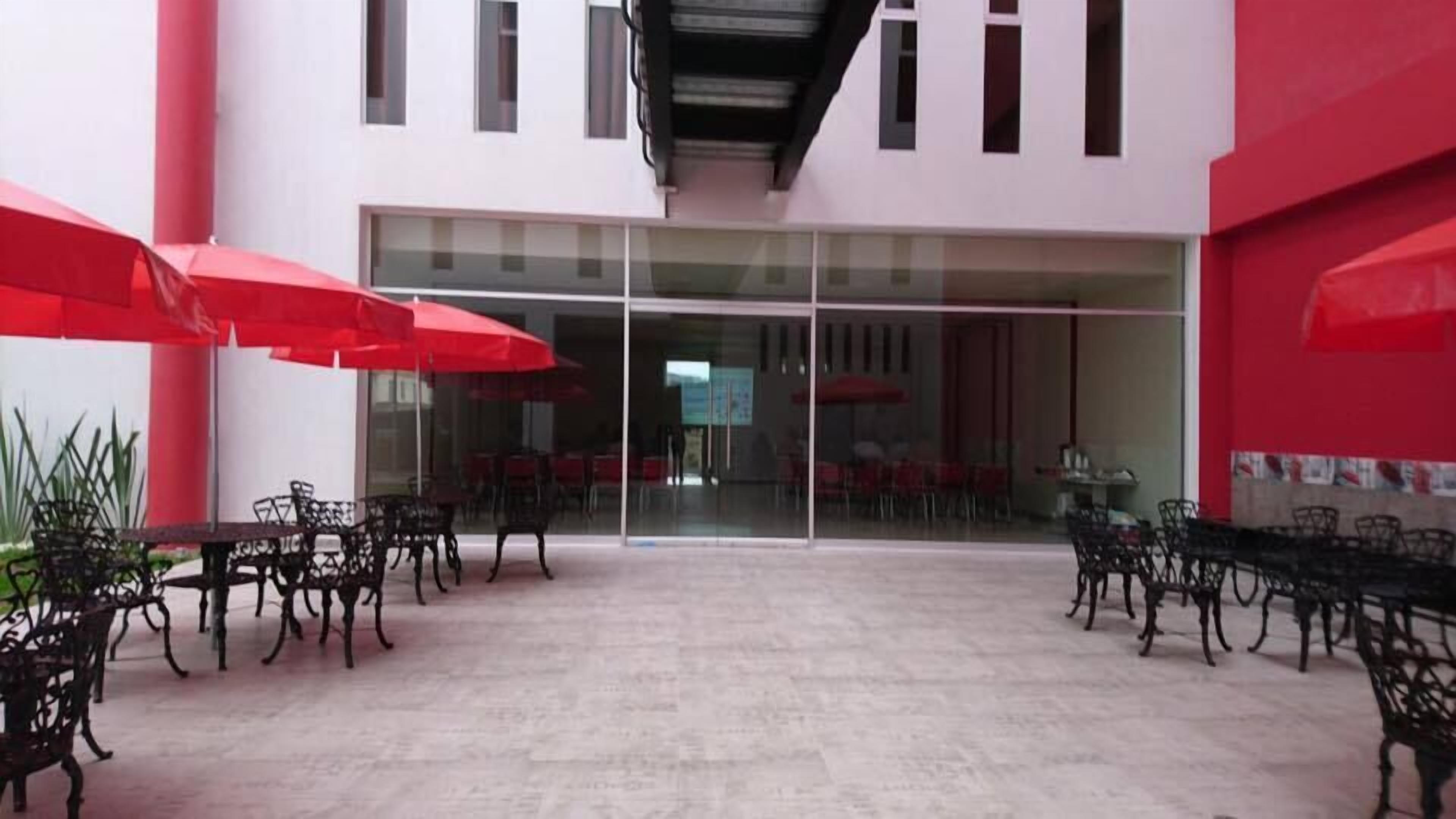 Un patio con mesas y sillas, sombrillas rojas y una entrada con puerta de vidrio.