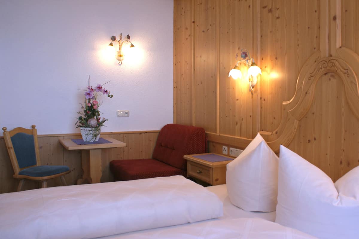 A hotel room with a wooden headboard, a bed with white linens, a red chair, a small table with a vase of flowers, and two wall-mounted lamps.