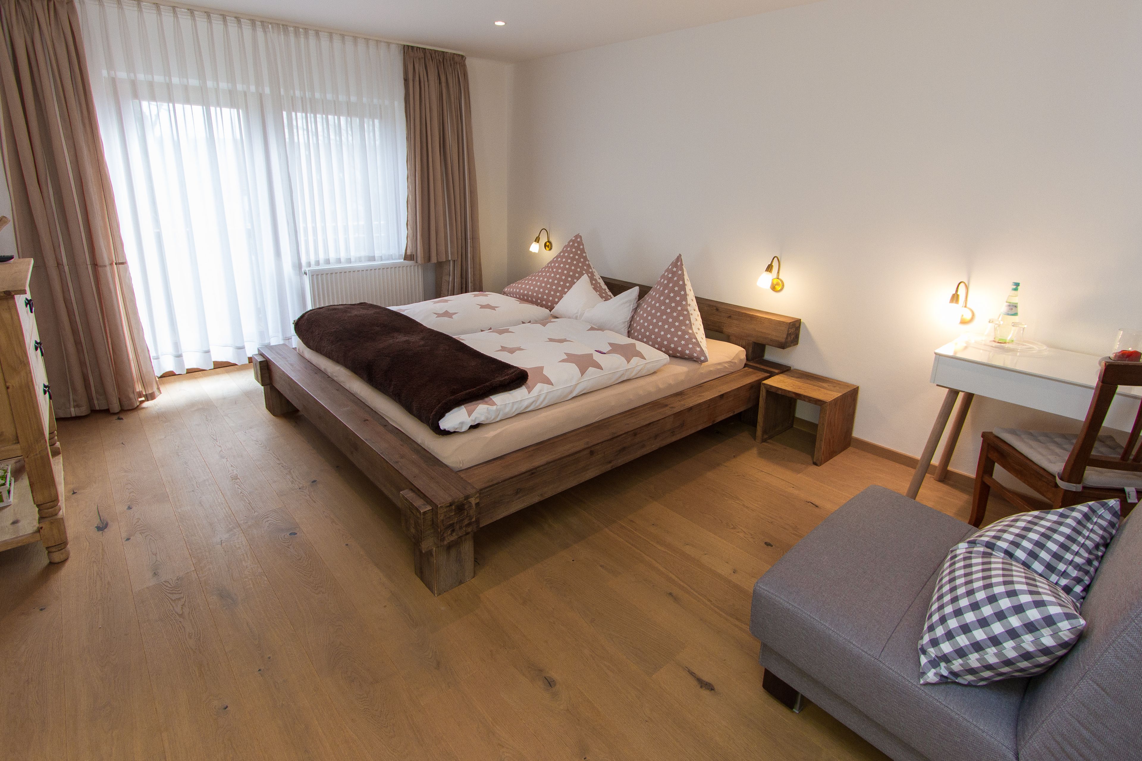 Ein Schlafzimmer mit einem hölzernen Bett, einem grauen Sofa, einem kleinen Tisch und einem Fenster mit Vorhängen.
