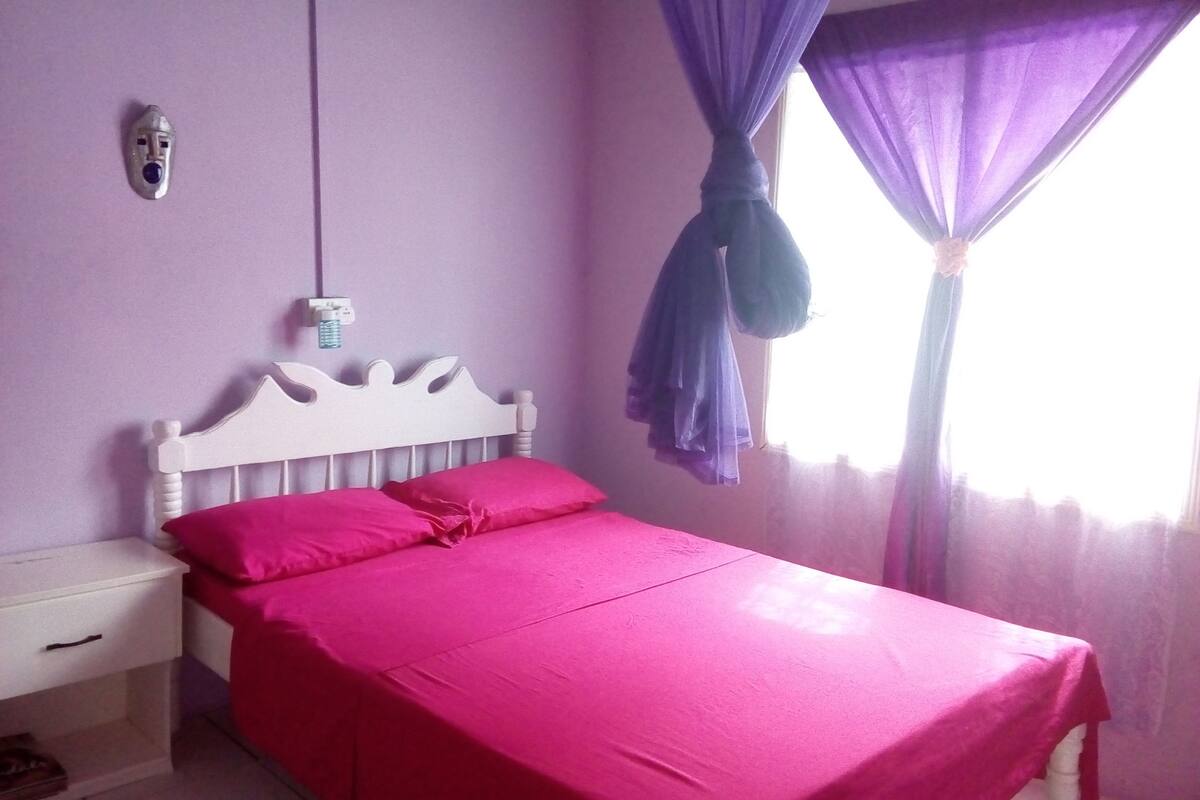 A bedroom with a white bed, pink bedding, a white nightstand, a wall-mounted light, and a window with purple curtains.