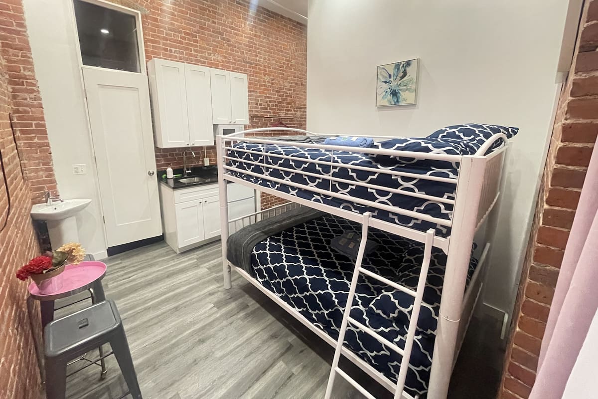 Una cama litera con una colcha estampada, un taburete con una bandeja rosa, un lavabo y un armario en una habitación con paredes de ladrillo y suelo de madera.