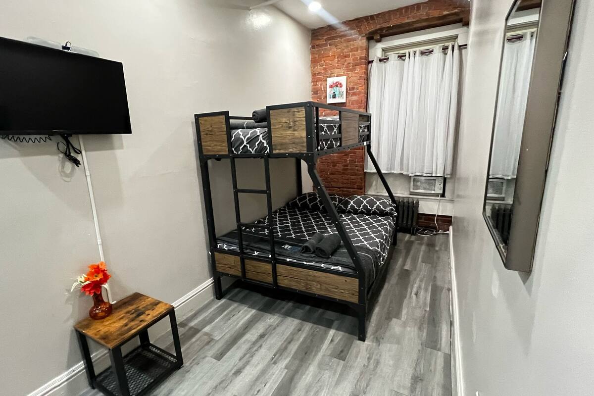 Una habitación pequeña con una cama individual, una mesa con un florero, un televisor de pantalla plana en la pared y una ventana con cortinas.