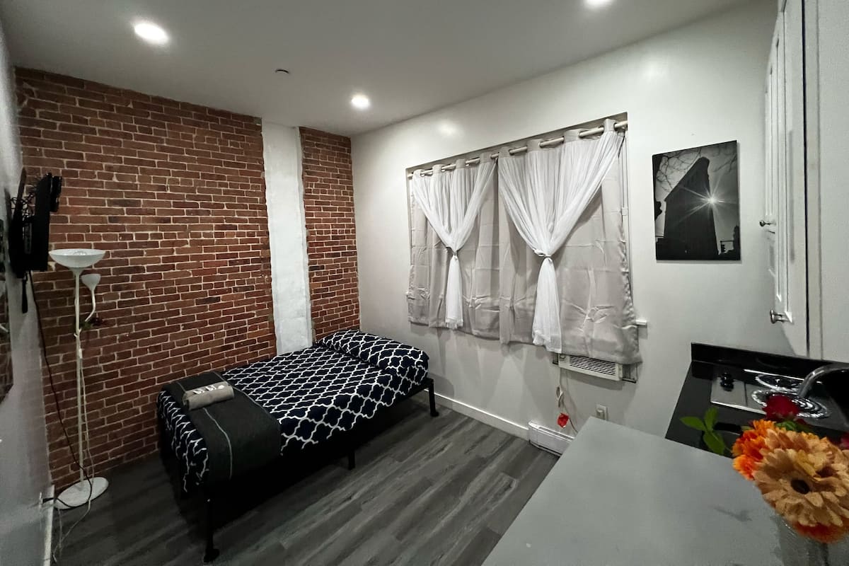 Un dormitorio con pared de ladrillo, una cama con colcha estampada, cortina blanca, una mesita con un florero y un cuadro en la pared.
