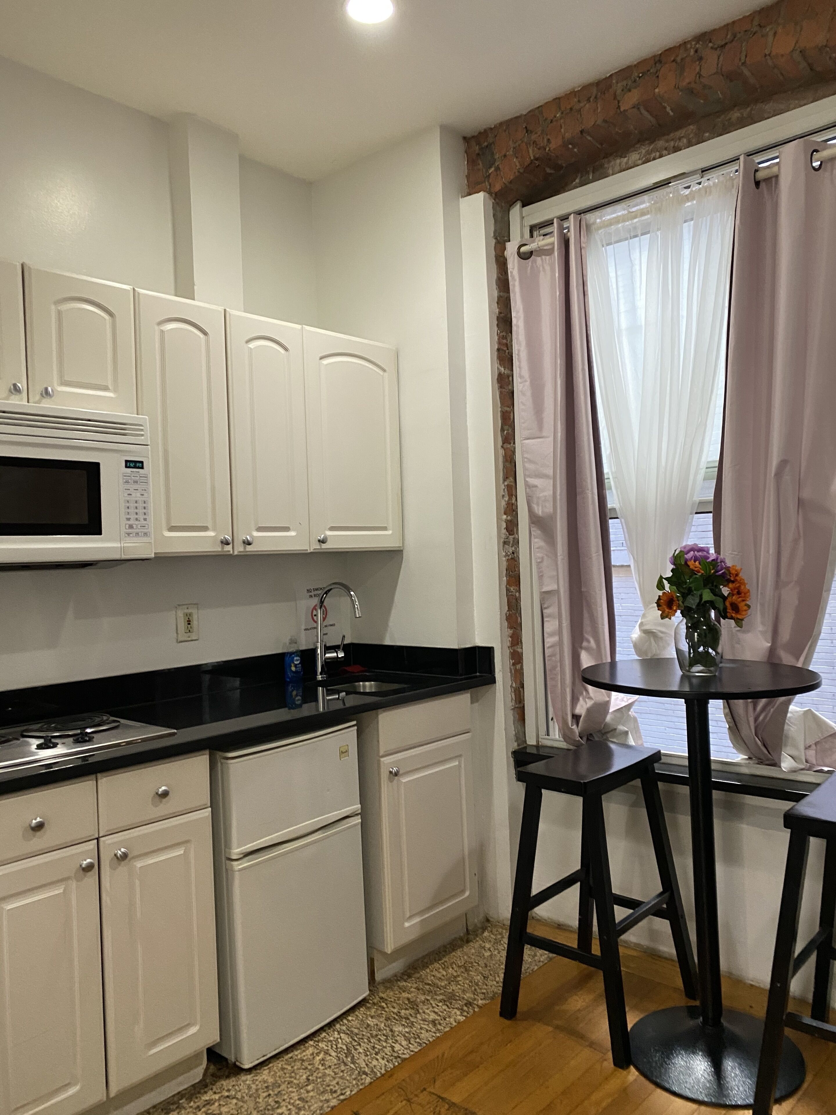 Cocina compacta con armarios blancos, microondas, lavamanos y una pequeña mesa redonda con un florero.