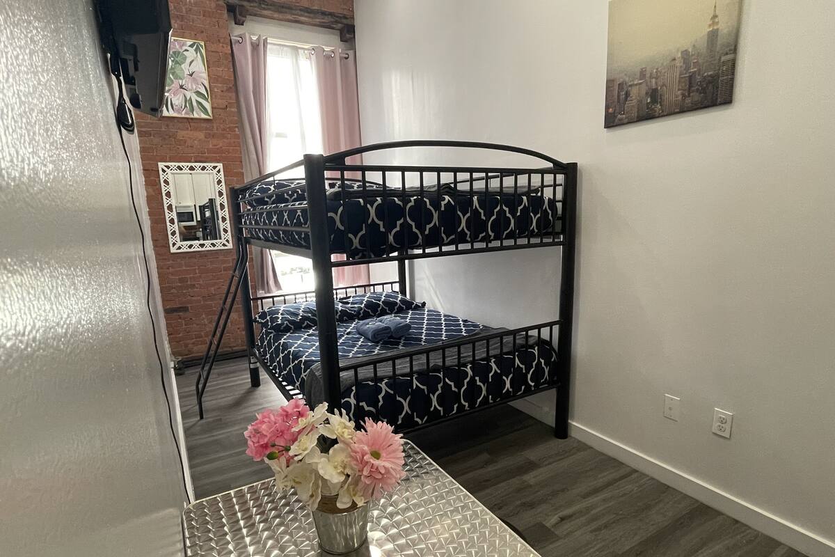 Una cama litera con una colcha estampada, una mesita con un florero y un cuadro de un paisaje urbano en la pared.