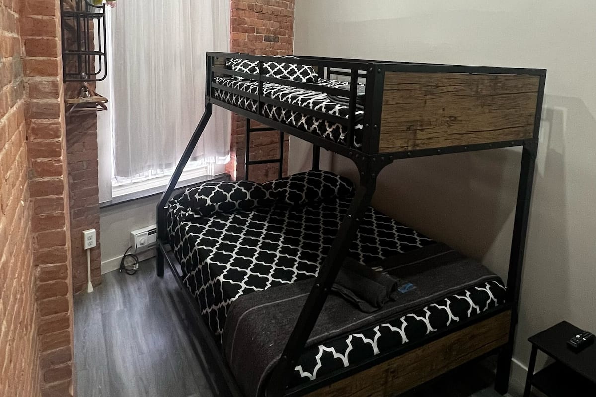 Una habitación con literas, paredes de ladrillo, una ventana con cortinas y una mesita.