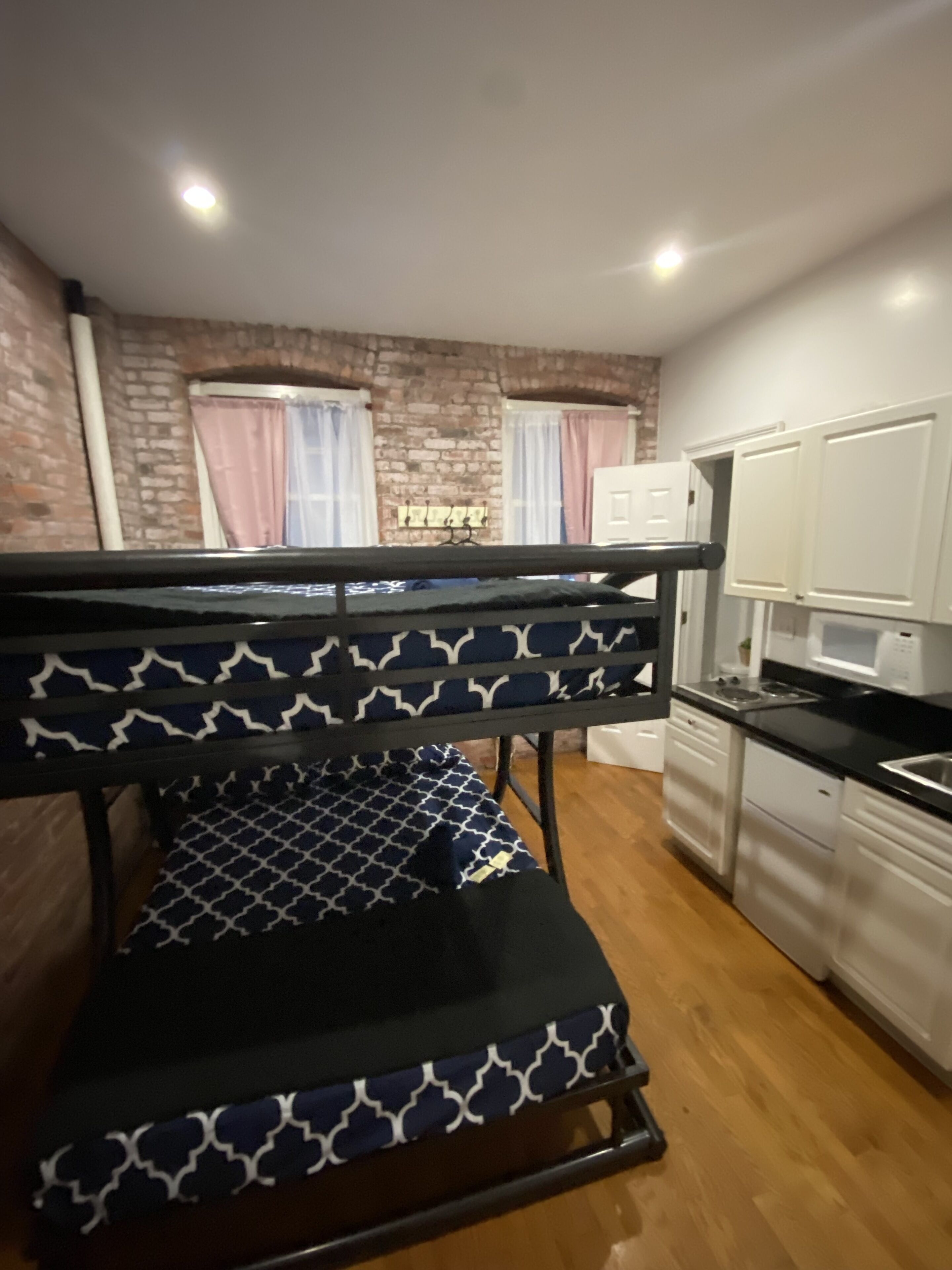 A bunk bed with a patterned bedspread, a kitchen area with white cabinets, and a brick wall in the background.